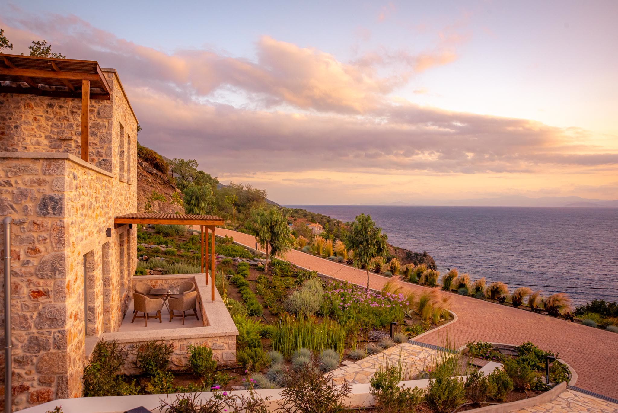 Stone-built suites at Sampatiki with a pergola terrace and planted slope above the Aegean at sunset