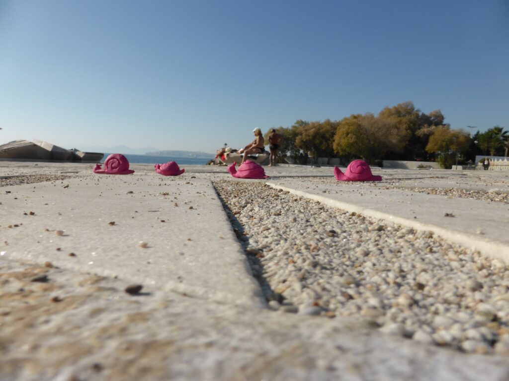 Pink ceramic snail sculptures placed along a gravel plaza in Athens